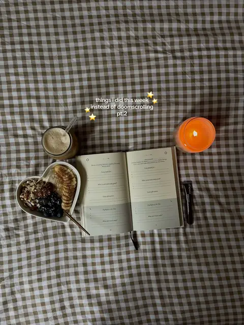 The image shows a scene on a bed with a brown and white checkered pattern. On the bed is an open journal with writing on the pages, a heart-shaped bowl filled with fruit and toppings, a glass of a brown beverage with a straw, and a lit orange candle. The overall composition is framed from a top-down perspective.