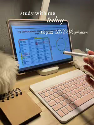 The image shows a well-lit workspace with a focus on a study session. In the center is a tablet displayed on a stand, showing a colorful diagram about \"DNA Replication\". A stylus rests on the screen, while a pink-keyed keyboard and a brown notepad with a spiral binding are placed below. The background is a softly lit desk, and the whole scene presents a clean, organized, and inviting study setup.