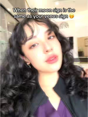 The cover image is a selfie-style shot featuring a young woman with dark, curly hair and a dark black jacket over a purple shirt. The background appears to be an indoor setting with lights and some greenery. Text overlayed on the image reads, 'When their moon sign is the same as your venus sign' with a smirking emoji. The woman is looking at the camera with pink lips and a subtle makeup.