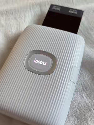 A close-up shot of a white Instax printer with a black piece of paper protruding from the top. The printer sits on a crinkled white surface. The Instax logo is illuminated on the front. The overall shot is well-lit, with a clean, minimalist aesthetic.