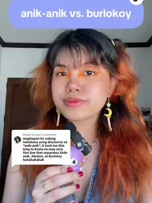 A woman with orange and black hair, wearing a denim top is holding a microphone and smiling. Above, there is text in a light blue box that says 'anik-anik vs. burlokoy.' To the left, there's a comment displayed from another user.