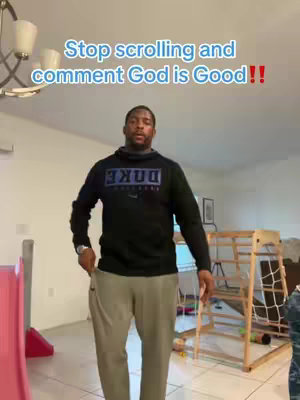 A man, dressed in casual clothing, is standing in a living room. The text overlay 'Stop scrolling and comment God is Good!!' is at the top of the frame.