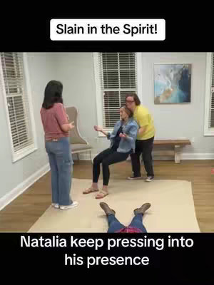 The image shows a room interior with three people standing and another lying on the floor suggesting a spiritual or religious context. Two people are holding a woman who seems to be fainting, while another woman stands nearby. The bottom of the image shows the feet of someone laying on the floor. The setting appears to be a home or a church, with neutral colors and basic furnishings.