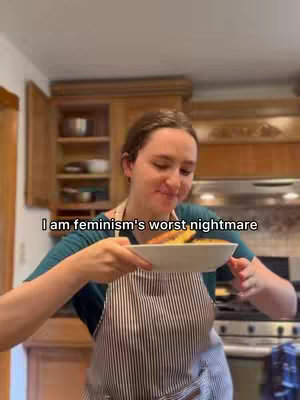 A woman in a kitchen holds a plate of food, looking at the camera. The background shows wooden cabinetry and a glimpse of a stovetop. The woman is wearing an apron.