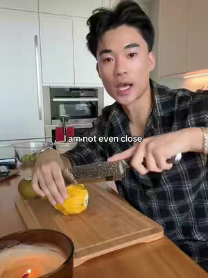 A young Asian man wearing a plaid shirt stands in a kitchen, holding a knife and cutting a mango on a wooden cutting board. Text overlay reads, 'I am not even close.' A lit candle and a bowl of fruit are in the foreground.