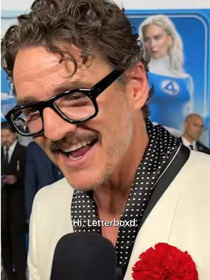 Close-up of a smiling man with glasses and a mustache, wearing a cream-colored blazer over a black and white polka-dotted scarf. He is set against a background that includes a poster for The Fantastic Four, with a woman in a costume visible. The man is holding a microphone and a red flower is pinned to his lapel. Text in the image reads, 'Hi, Letterboxd'.