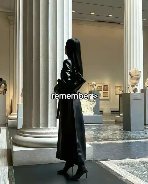 A woman in a long black dress and heels stands in a museum, looking at sculptures and exhibits. Text overlayed reads 'remember >'.