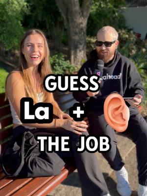 A cover image showing a woman and a man seated outdoors on a bench. The woman is on the left, smiling expansively. The man sits to her right, wearing sunglasses and holding a microphone. Overlaid on them is a text overlay that reads "GUESS La + THE JOB" using a white font on a solid black background. In the background, there is greenery and a tree.