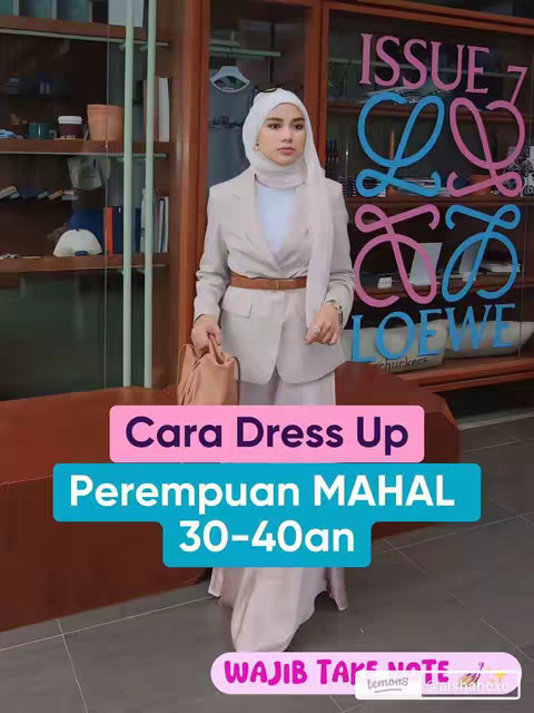 A woman in her 30s or 40s, wearing a cream-colored blazer and pants with a white headscarf and a brown belt, poses in front of a Loewe storefront. Text overlays in pink and blue on a pink background highlight the video's theme: 'Cara Dress Up Perempuan MAHAL 30-40an.' The overall composition is stylish and fashionable.