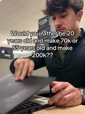 A man is shown in an office environment, holding open a laptop which obscures his lower body. The overlay text poses a question about choosing between earning 70k at age 20 or 200k at age 55.
