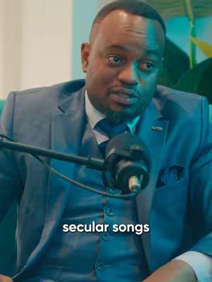 A close-up shot features a Black man wearing a suit, sitting in a studio setting with a microphone in front of him. The background is subtly lit with blue and green hues.