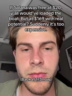 A selfie-style video shows a man from the chest up, against a blurred background. He is looking toward the right side of frame with a neutral expression. Text overlays the image in white. Above the man's head, text poses a question relating to the value of Solana. At the bottom of the frame, the text includes a comment on the current price of Solana. The man has short brown hair, a short beard, and a light complexion .