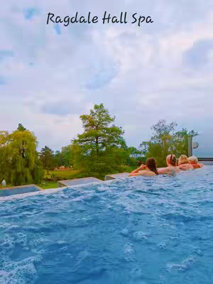 The image shows an outdoor spa scene with a heated rooftop infinity pool overlooking a green landscape. Above the pool, a cloudy sky fills the frame. The text "Ragdale Hall Spa" is overlaid on the top. Several people are enjoying the pool, with their heads visible above the water.