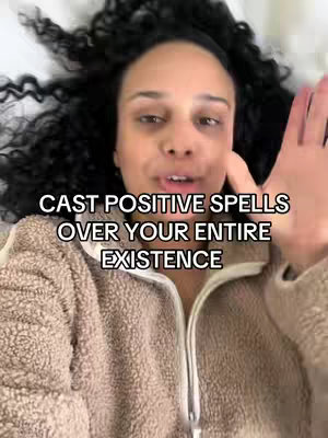 A woman with dark, curly hair is lying on a bed. She is wearing a brown, zipped up jacket and has one hand raised towards the camera. Text overlayed onto the image at the top reads "CAST POSITIVE SPELLS OVER YOUR ENTIRE EXISTENCE".