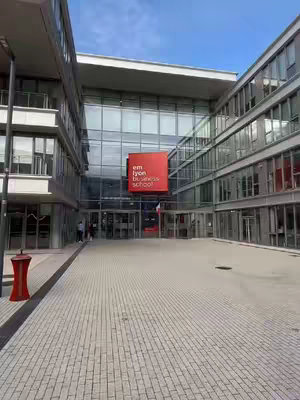 An exterior shot of a modern building with a glass facade. The entrance is visible, with a red square logo of 'emlyon business school' above the doors. The foreground is a paved area. The overall composition is symmetrical, with the building filling most of the frame.
