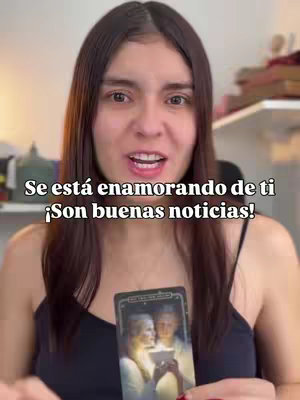 A woman with long dark hair looks directly at the camera, smiling slightly. The background is out of focus but appears to be a room with neutral colors. She is holding a tarot card with figures depicted on it, and has a text overlay on top that says, "Se está enamorando de ti ¡Son buenas noticias!"