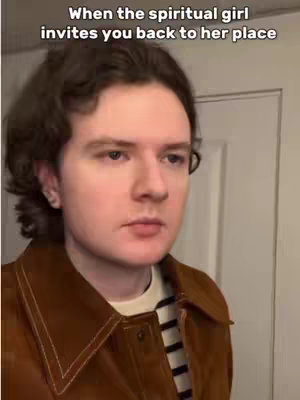 A medium shot shows a young man indoors, gazing slightly down and to the left. He's wearing a brown, corduroy-like jacket over a striped black and white shirt. The background is blurred, suggesting a room interior with a white door on the right.