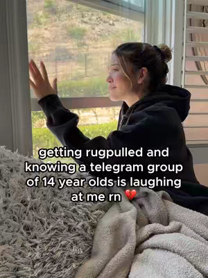 A young woman is sitting by a window, looking out. She has her hair up in a bun. She is wearing a black hoodie and has her hand up. The background shows a blurry outdoor scene.