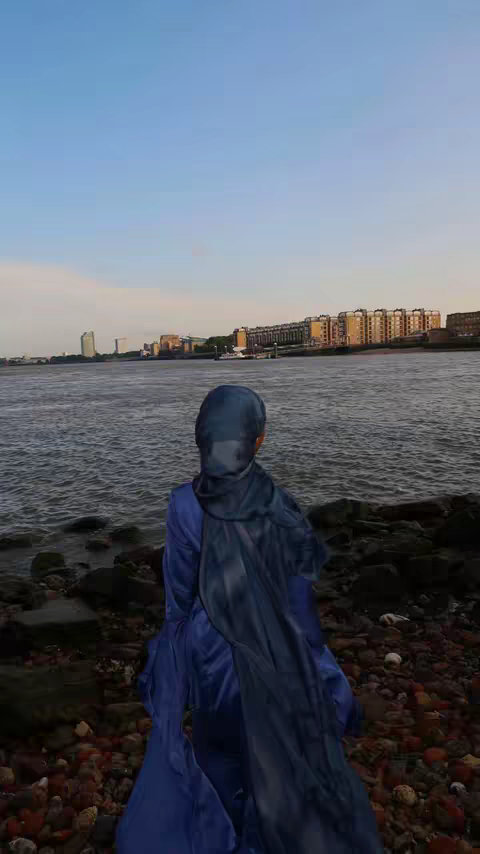 A person wearing a dark blue hijab and dress stands with their back to the camera, looking out at the water. The sea takes up about half the frame. Buildings line the horizon in the distance under a slightly cloudy sky.