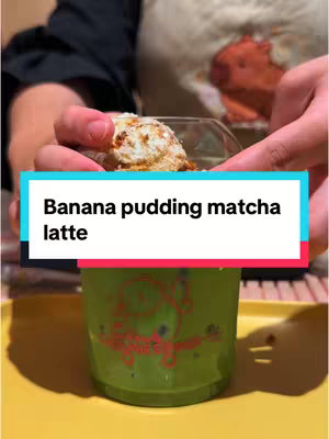 A person is shown holding a clear glass cup with a matcha drink with what appears to be banana pudding and a topping within the cup. The cup sits on a yellow tray. The person's hands are visible, and a teal-colored box with white text overlayed on it reads: 'Banana pudding matcha latte'. Behind the person, a portion of a person in an apron is seen.