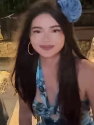 A woman with dark hair and a blue floral hair accessory smiles directly at the camera. She wears a halter-style top with a blue and white pattern. Gold hoop earrings are visible. The background is softly blurred.