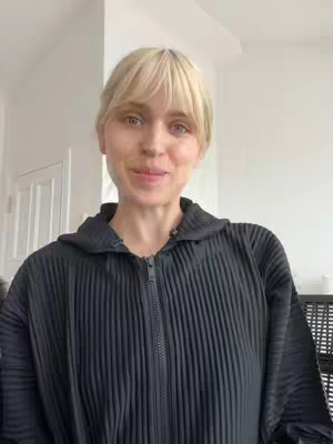 The image shows a woman in a black hooded jacket. She has blonde hair with bangs and fair skin. She is looking directly at the camera with a slight smile.