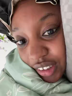A close-up shot of a person's face, mostly obscured by a green, floral-patterned sheet. The person is looking towards the camera with dark eyes, a slightly open mouth, and a soft, inviting expression. A black satin bonnet, with a white trim, frames some of the hair and the head. The overall tone is intimate and casual.