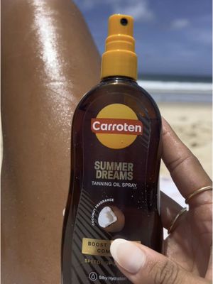 The image shows a person's leg next to the beach. A bottle of Carroten Summer Dreams tanning oil spray is held in front of the leg. The background is a blurred beach scene with the blue sky visible. The person is holding the tan oil bottle with a hand that has nice nails with a gold ring on one of the fingers. The image has a summery feel.