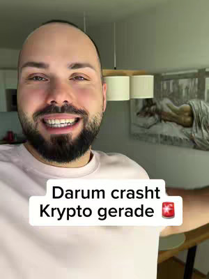 A smiling man with a beard is the central figure in the image. He is wearing a light pink shirt. The background shows a kitchen with a chandelier. The image has a text overlay at the bottom with the words 'Darum crasht' on the first line, 'Krypto gerade' second line, and an icon of a red emergency light.