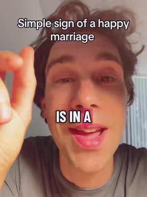 A close-up shot of a young man's face with text overlays. The text reads 'Simple sign of a happy marriage', with the words 'IS IN A' overlaid in a white box beneath it. The man appears to be speaking, with his mouth open, his skin is fair, and has dark hair. He is wearing a grey shirt.