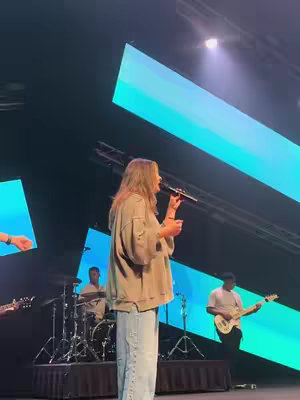A woman with long hair stands center stage singing into a microphone. She faces towards the side of the frame, and is wearing casual clothes. There are band members in the background, consisting of a drummer and two guitarists. The stage is lit with blue and purple lights. There are stage lights above the performers.
