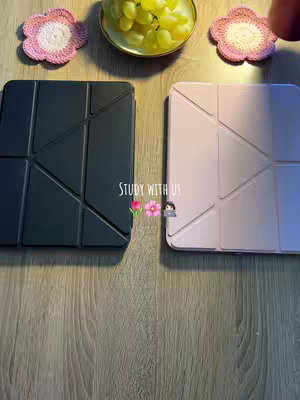 The image is a top-down view of a wooden table. On the table are two iPad cases, one black and one pale pink, arranged side-by-side. A plate with green grapes is placed above the cases. Two pink flower-shaped crocheted objects are on either side of the plate. The text overlay "STUDY WITH US" is written in a sans-serif font.