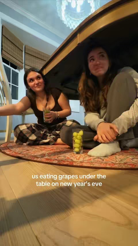 Two young women sit on a patterned rug under a table, with one holding a green drink. A glass of grapes sits nearby. The setting is a room with wooden floors and a chandelier.