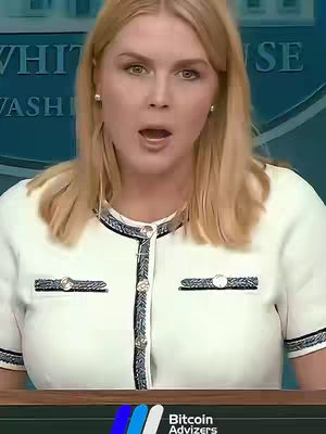 A blonde woman with widened eyes and an open mouth is positioned center frame, appearing surprised. She wears a white jacket with black trim and has a pearl earring in each ear. The scene appears to be set in front of a backdrop with a blurred inscription that appears to be White House. The bottom of the frame features a logo for "Bitcoin Advizers".