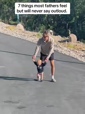 A man and a toddler are walking on a gray surface, likely a driveway or path. The man, dressed in a light-colored long-sleeve shirt and dark shorts, is holding the toddler's hands to help them walk. The toddler is wearing a black long-sleeved shirt and light-colored shorts The setting appears to be outdoors, as suggested by the trees and dry foliage in the background.