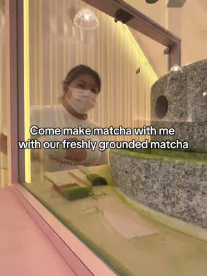 The image shows a person behind a counter, possibly in a cafe, wearing a face mask. The person is facing the camera, slightly out of focus. In the foreground is a counter with matcha-related items like green matcha powder, and a grinding mechanism.