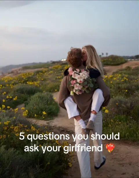 A couple is embraced in a kiss on a road, the man holding the woman as they enjoy the outdoors. They are surrounded by a field of yellow flowers with a background that indicates dusk. The text overlay states: "5 questions you should ask your girlfriend".