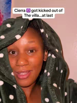 A close-up shot of a Black woman with dark skin, gazing directly at the camera. She's wearing a hooded robe with a dark green fabric covered in white polka dots. Text overlay at the top states, 'Cierra ♎ got kicked out of The villa...at last'. The background is a blurred pinkish hue, softly illuminating the subject.