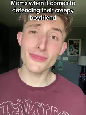 A young man with light skin and short, styled hair is centered in the frame. He is wearing a maroon t-shirt. The background is a slightly blurred, dark setting that appears to be indoors, possibly a room. The man’s facial expression is soft, with a slight, subtle smile. Above him, the text overlay reads, 'Moms when it comes to defending their creepy boyfriend'.