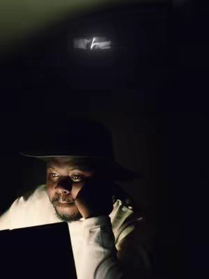A dimly lit image featuring a dark-skinned man with a beard wearing a black hat. He is leaning towards a laptop, with the screen illuminating his face. Above him, a faint light source casts a glow on the ceiling. The overall composition is dark and moody.