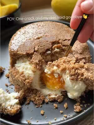 A close-up shot of a no-bake mango cheesecake sliced to reveal the layers. The cake has a biscuit base, a creamy white filling, and a vibrant orange mango layer. A hand holding a utensil is in the frame, about to take a bite. The cake sits on a dark plate, with a mango fruit in the background with the text 'POV: One crack and my diet said goodbye'.