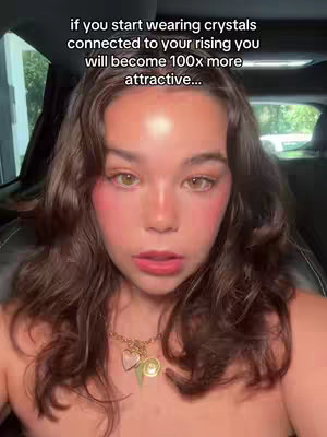 A selfie-style shot featuring a young woman with light skin, brown wavy hair, and a slightly startled expression. She has rosy blush on her cheeks and a subtle dark lip color. She's wearing a necklace with a heart pendant, several charms, and a gold chain. A text overlay, written in a white san serif font, is positioned at the top, and the background appears to be of a car interior. 