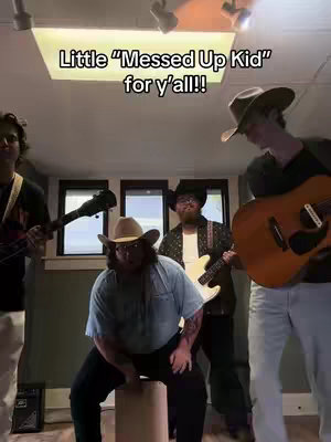 A group of four men, dressed in cowboy hats and casual wear, are captured in an indoor setting. The one in the center is sitting on a rounded object, surrounded by the others who are holding instruments. The overall lighting is warm, and the composition includes text overlay at the top.