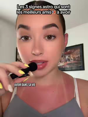 A young woman with fair skin and dark hair is holding a small black microphone in front of her mouth while looking directly at the viewer. She wears a gray tank top, and her shoulders and chest appear sunburned with the skin reddened. Above her, a text overlay reads, "Les 3 signes astro qui sont les meilleurs amis à avoir," framed in black. Below her, the text "AVOIR DANS SA VIE" is present. The background features an out-of-focus interior with framed artwork.