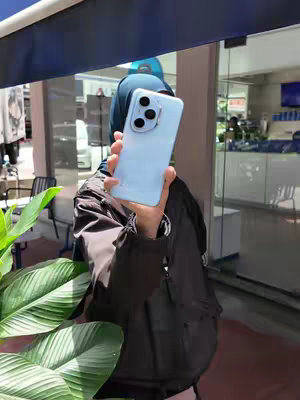 A person is holding up a light-blue smartphone with a triple-lens camera. The person's face is covered by a hijab. The background includes greenery, an outdoor setting with a glass door and a blue awning.