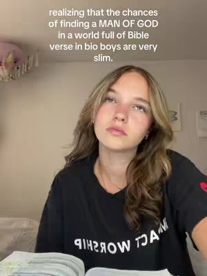 A young woman with long, wavy light brown hair and a thoughtful expression is looking upwards. She's wearing a black t-shirt with white text and is sitting with a partially open book in her lap. The background is a plain wall. Overlayed text in white, is visible at the top of the image.