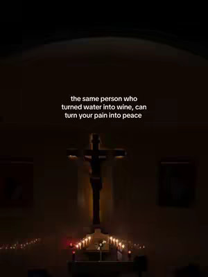 The image is a dimly lit interior shot, likely a church or chapel. A large crucifix is centrally placed, with a figure of Jesus on it. Below the cross, a simple altar or ledge holds lit candles. In the foreground, text overlaid on the image conveys a religious message, using a clean, sans-serif font. The overall mood is reverent and solemn, with a focus on faith and spirituality.
