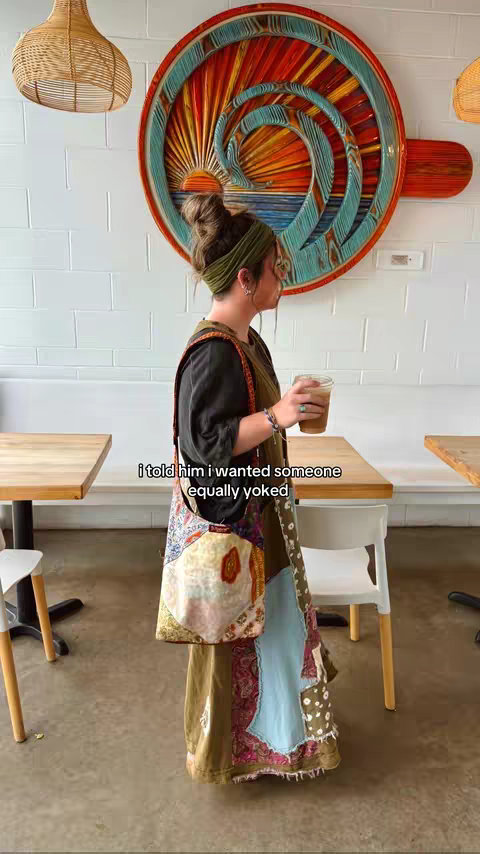 A woman stands in a cafe, holding a drink. The background features a decorative wall hanging and tables. Text overlay on the image reads: "i told him i wanted someone equally yoked".