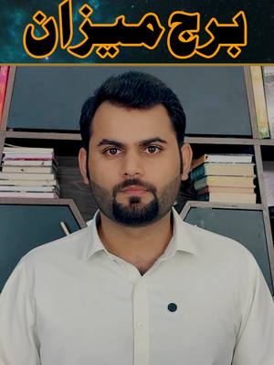 A medium shot of a man facing the camera, set against a dark background with bookshelves in the mid-ground. The man has short black hair and a neatly trimmed beard. He wears a white button-down shirt, and has a focused expression.