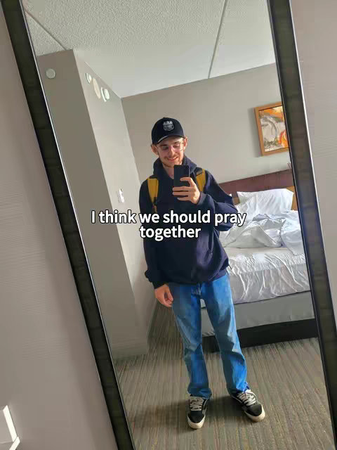 A selfie taken in a hotel room setting. The main subject, a young man, is reflected in a large mirror. He is wearing a dark blue hoodie, blue jeans, a backward black cap, and glasses. He is holding a smartphone, and a yellow backpack strap is visible over his shoulder. The text overlay in the center of the image reads, 'I think we should pray together'. The composition includes a section of the room consisting of a bed with pillows under the mirror.